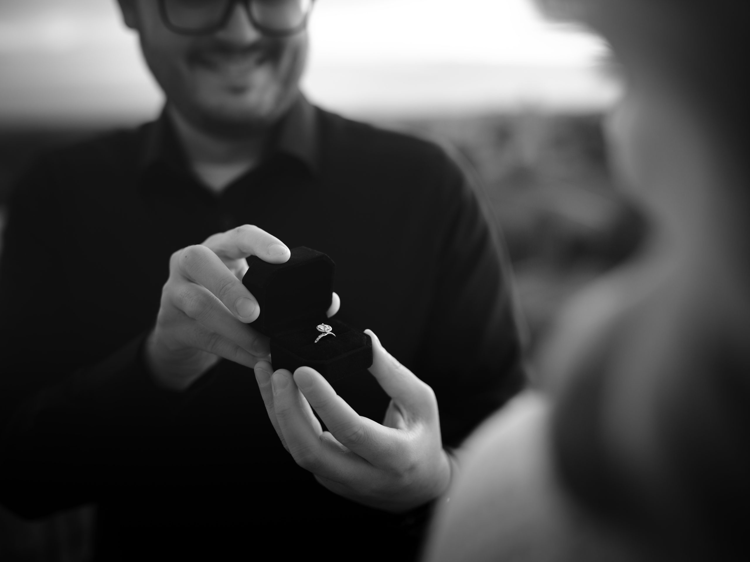 Black and white image of a man opening a small ring box to present an engagement ring, capturing a meaningful life moment in the ENVISTABLACK Better Together photography campaign.