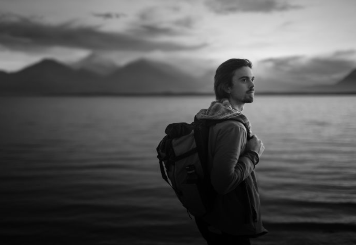 Black and white portrait of a young man with a backpack standing by a lake, looking toward the horizon, representing ambition and possibility in the ENVISTABLACK photography campaign.
