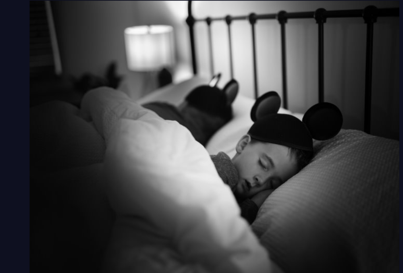 Black and white image of a person sleeping peacefully in bed while wearing playful mouse‑ear headwear, conveying comfort, rest, and everyday life moments.